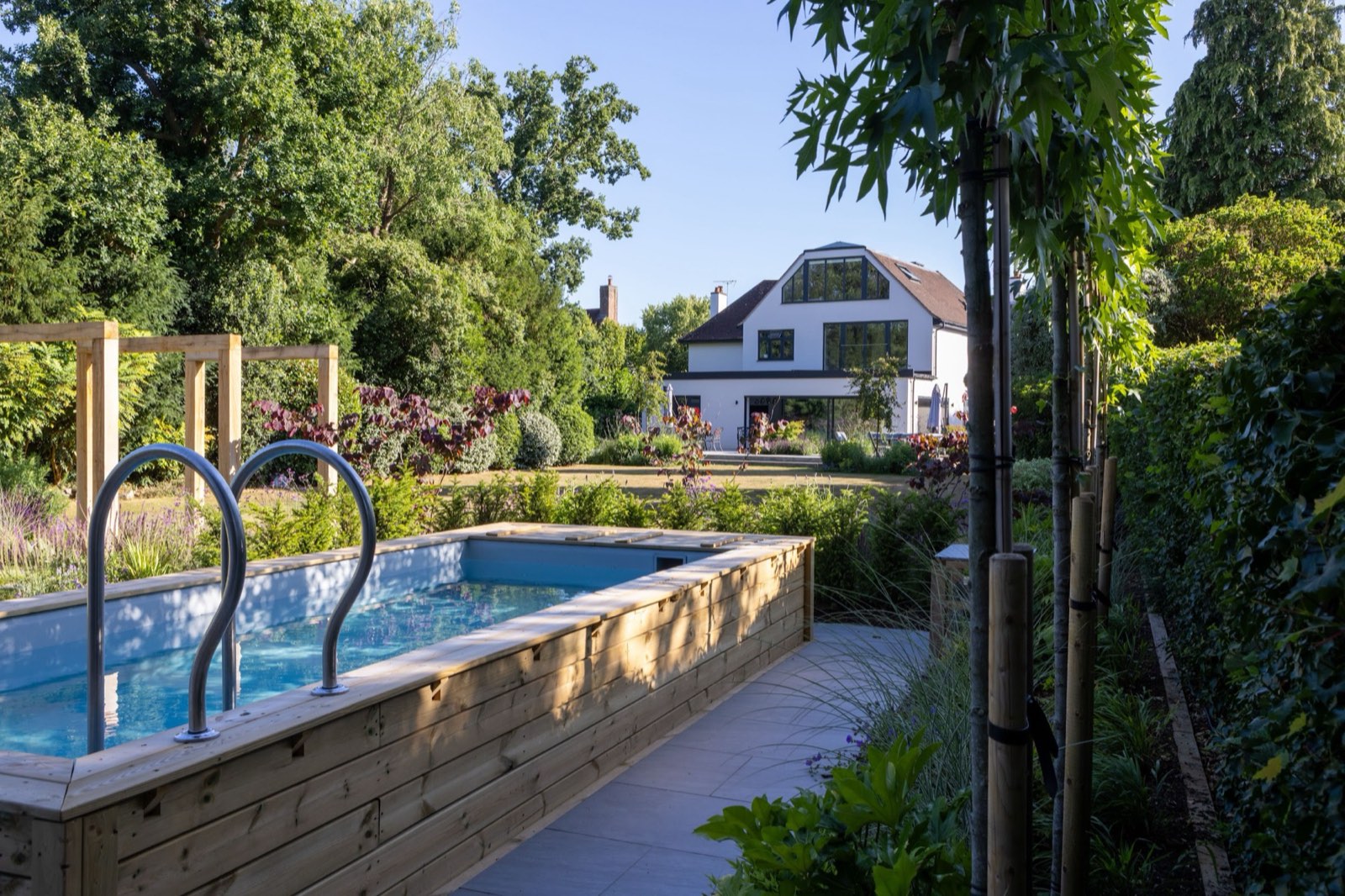 A raised timber-clad pool set into planting, with the house framed by trees beyond in evening light.